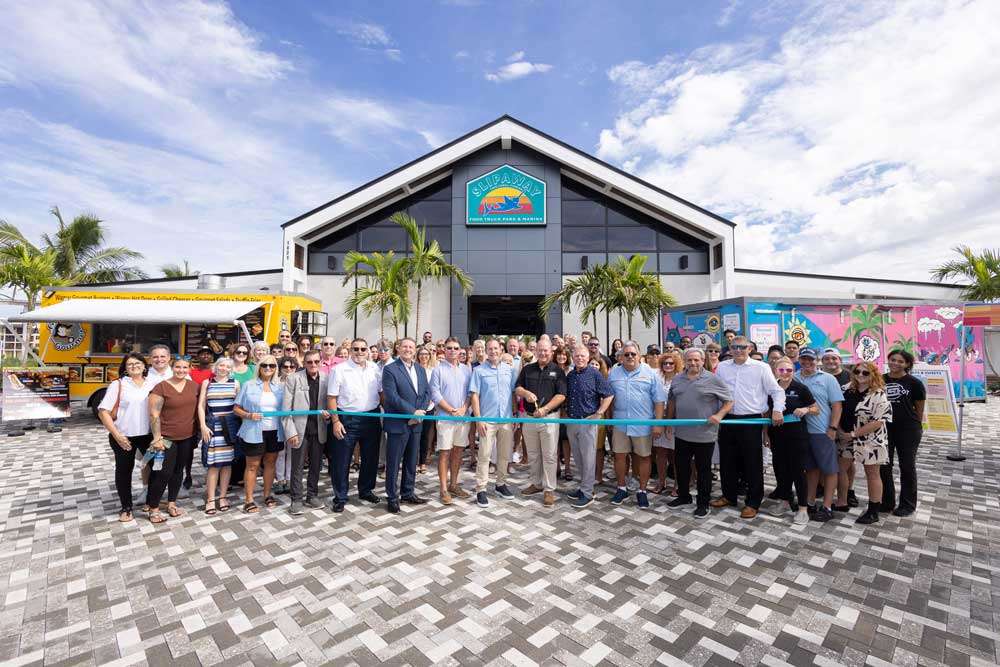 A group of people stands in front of a building, holding a blue ribbon, celebrating a special event or opening.