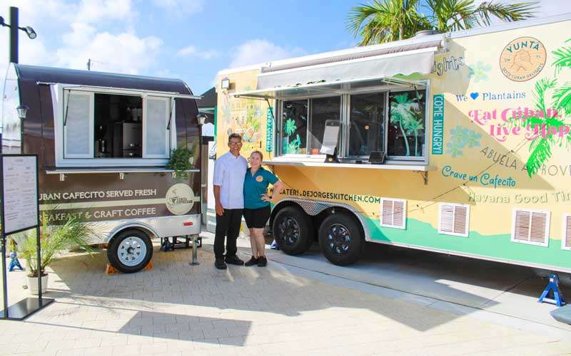 A man and woman stand smiling between a small coffee trailer and a large food truck on a sunny day, both trucks featuring Cuban-themed branding and designs.