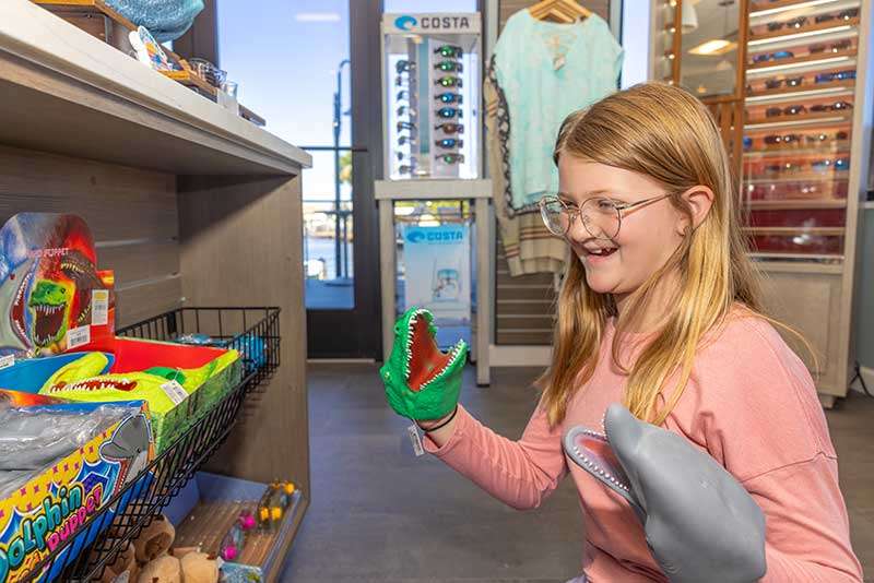 A girl wearing glasses smiles while playing with toy animal hand puppets in a store aisle filled with various toys and sunglasses.