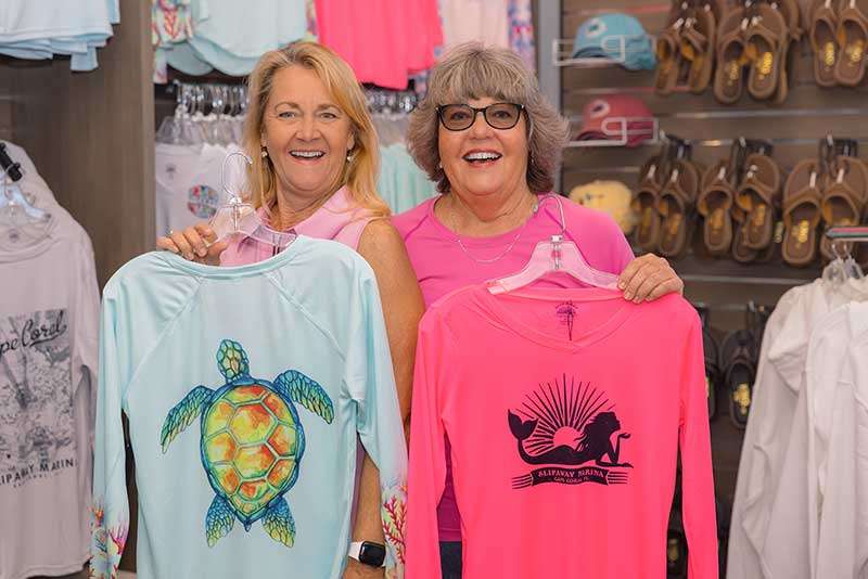 Two women standing in a clothing store hold up long-sleeve shirts, one with a sea turtle design and the other with a Saturday Island graphic.
