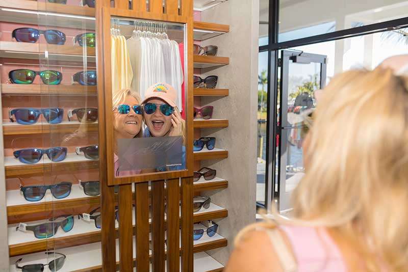 Two women wearing sunglasses smile and take a selfie in front of a mirror in a store display with multiple sunglasses.