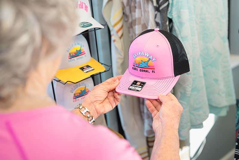 A person holds a pink "Slipaway Cape Coral, FL" trucker hat in a store, with more hats displayed in the background.