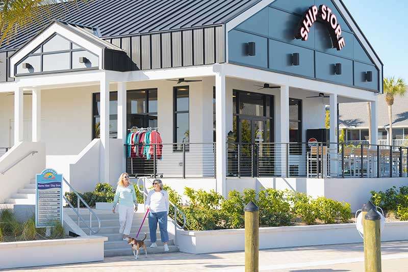 Two women walk a dog past a white ship store with outdoor seating and a clothing rack visible near the entrance on a sunny day.