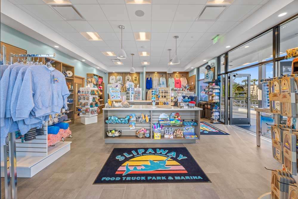 Gift shop interior with neatly arranged clothing, souvenirs, and accessories on shelves and racks; large windows to the right; “Slipaway” logo mat on the floor near the entrance.